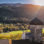 La tour Montmayeur, en pierre, parmi des maisons, avec des rayons de soleil sur une foret et des montagnes en arrière-plan.