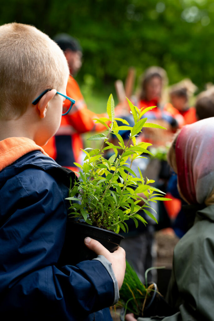 Un enfant de dos tient dans la main une plante en pot remise par l'un des agents des espaces verts de la commune présent en flou dans le fond de l'image