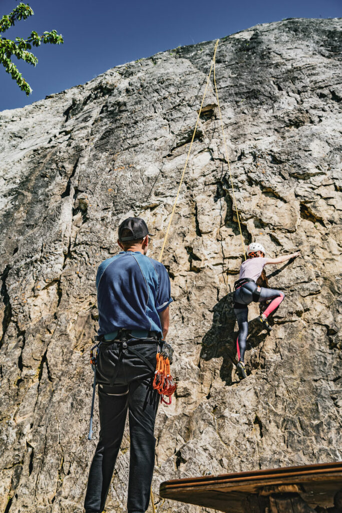 Un homme de dos assure grâce à sa corde une jeune fille avec un casque grimpant l'un des itinéraires rocher du site d'escalade de Villette
