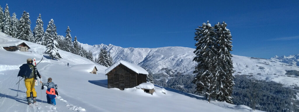 Un père de dos et avec des skis dans le dos tient la main de sa petite fille. Ils remontent la piste de le décor enneigé de l'espace nordique de Prachanier à Granier sous un beau ciel bleu