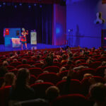 Une femme déguisée en ménagère et seule sur une scène représentant une cuisine fait face à des enfants de dos sur les fauteuils rouge de la salle de spectacle d'Aime-la-Plagne
