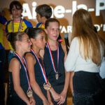 Des jeunes sportives en survêtement noir, médailles au cou, se tiennent sur scène lors de la cérémonie de la Soirée des Champions à Aime-la-Plagne. Elles parlent à un femme de dos leur présentant un micro.