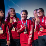 Des jeunes footballeuses en survêtement rouge, médailles au cou, se tiennent sur scène lors de la cérémonie de la Soirée des Champions à Aime-la-Plagne. L’une d’elles, souriante, tient un micro pendant que ses coéquipières l’applaudissent chaleureusement.